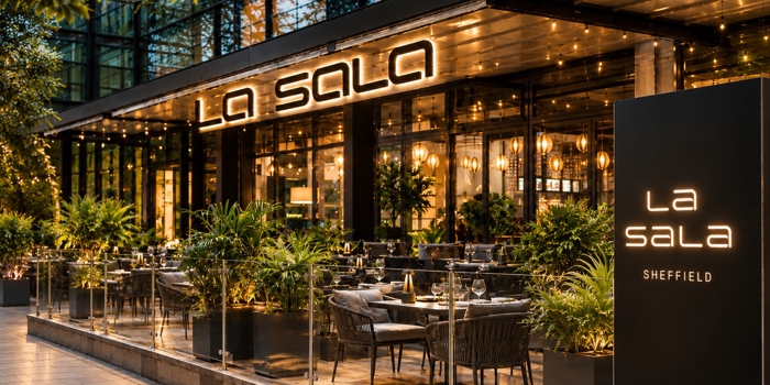 La Sala Sheffield restaurant exterior at dusk, featuring a stylish outdoor terrace with dining tables, lush planting, warm lighting and illuminated signage. Floor-to-ceiling windows reveal a contemporary interior, creating an inviting atmosphere
