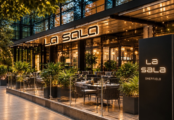 La Sala Sheffield restaurant exterior at dusk, featuring a stylish outdoor terrace with dining tables, lush planting, warm lighting and illuminated signage. Floor-to-ceiling windows reveal a contemporary interior, creating an inviting atmosphere