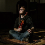 A person sat cross legged on the floor with a violin and bow in their lap.