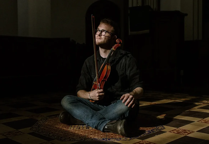 A person sat cross legged on the floor with a violin and bow in their lap.