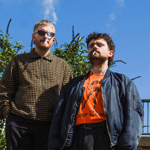 Two people stand outdoors beneath a clear blue sky, framed by leafy shrubs and a metal railing in the background. One wears sunglasses and a brown checked jacket, while the other wears a dark bomber jacket over a bright orange graphic T-shirt. Both are smoking cigarettes. The low-angle photograph creates a bold, stylised promotional portrait with strong sunlight and contrasting colours.