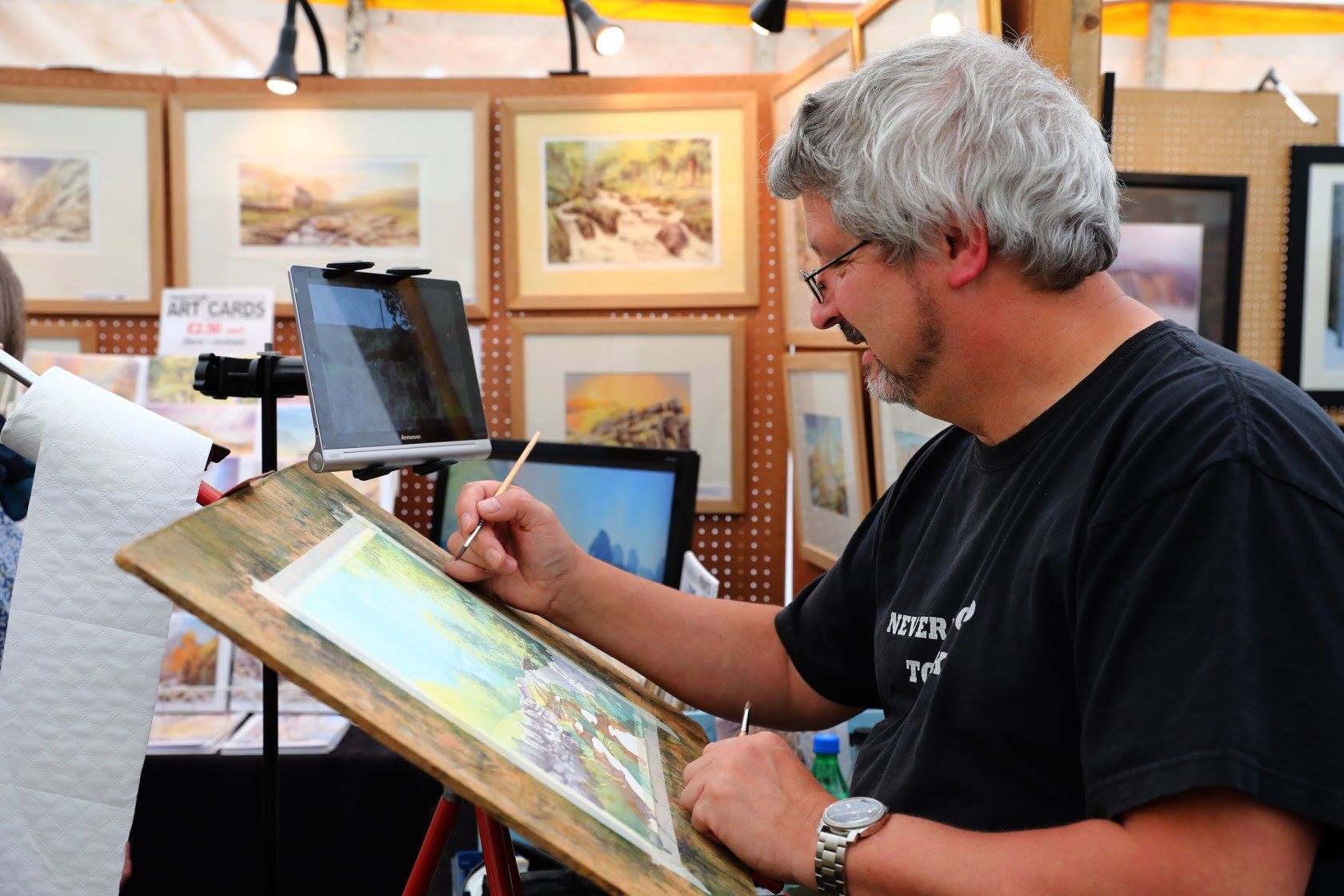  An artist is seated at a stall painting a landscape scene on a wooden board using a fine brush. The workspace includes a roll of paper towels, a tablet mounted on a stand, and several framed artworks displayed on pegboard panels in the background. The paintings feature natural scenery in soft, muted tones. A small sign on the left reads “Art Cards,” suggesting items for sale. The setting appears to be an art fair or exhibition.