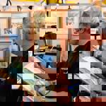  An artist is seated at a stall painting a landscape scene on a wooden board using a fine brush. The workspace includes a roll of paper towels, a tablet mounted on a stand, and several framed artworks displayed on pegboard panels in the background. The paintings feature natural scenery in soft, muted tones. A small sign on the left reads “Art Cards,” suggesting items for sale. The setting appears to be an art fair or exhibition.