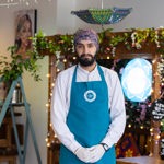 Person wearing a teal apron and white gloves stands inside a decorated shop or café. The background features hanging string lights, colorful cushions, framed artwork, and a wooden structure adorned with flowers and greenery. A round illuminated sign and a chalkboard with text are also visible.