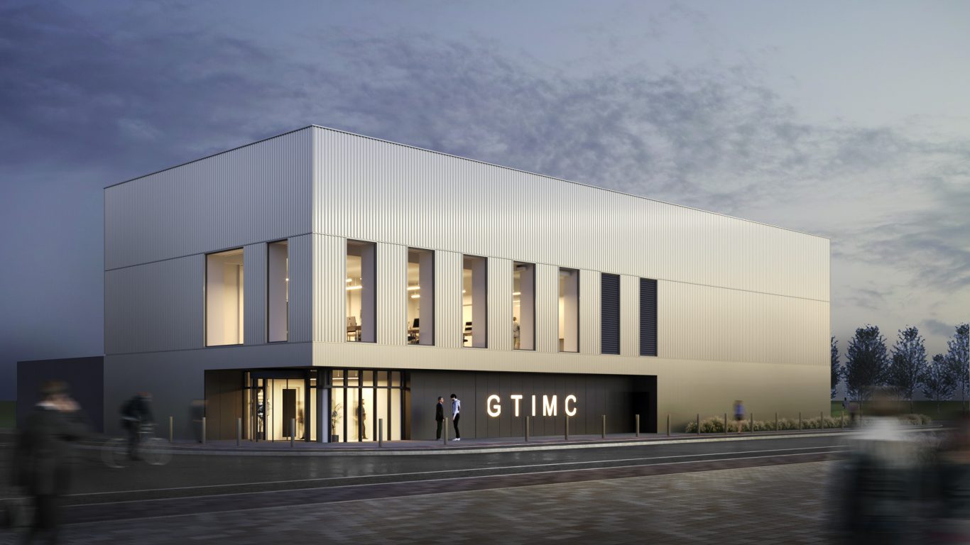 The exterior of the Gene Therapy Centre building. Modern two-story building with a sleek, minimalist design featuring vertical metal panels and large rectangular windows. The structure is illuminated from inside, creating a warm glow against the twilight sky. The entrance has glass doors and signage reading “GTIMC.” A few people are visible near the entrance, and blurred motion suggests activity in the foreground.
