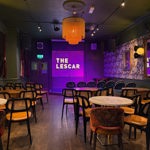 A cosy performance room at The Lescar with rows of chairs facing a small stage lit in purple, displaying the sign “Live at The Lescar,” and a large amber fringe lampshade hanging from the ceiling.