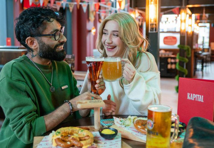 Two people are sitting at a table in a beer hall, clinking glasses of beer in a toast. One glass is a tall, dark beer and the other is a large mug of pale beer. The table has wooden boards with soft pretzels, mustard dip, and branded paper. A red menu with “KAPITAL” is visible, and the background features colourful bunting, warm lighting, and a modern interior with booths and bar seating.