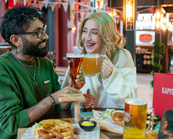 Two people are sitting at a table in a beer hall, clinking glasses of beer in a toast. One glass is a tall, dark beer and the other is a large mug of pale beer. The table has wooden boards with soft pretzels, mustard dip, and branded paper. A red menu with “KAPITAL” is visible, and the background features colourful bunting, warm lighting, and a modern interior with booths and bar seating.