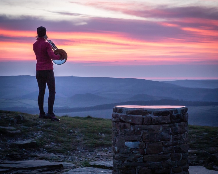 A person standing on a rocky hilltop at sunrise, holding a brass instrument while looking toward the horizon. The sky is filled with vibrant shades of pink, orange, and purple, casting a soft glow over the rolling hills in the distance. In the foreground, a circular stone structure sits on the ground, adding texture to the natural landscape.