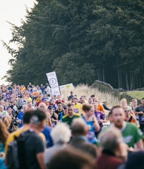 A large crowd is watching and cheering on runners running on a road. In the background is a large wood.