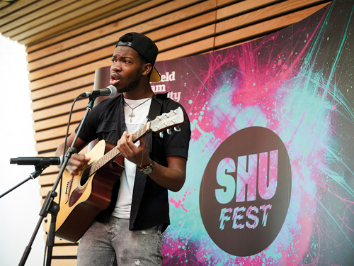 A person is performing on stage, playing an acoustic guitar and singing into a microphone. The backdrop features a colorful design with the text “SHU FEST” and “Sheffield Hallam University.” The setting appears to be an indoor event space with wooden panelling and stage lighting.