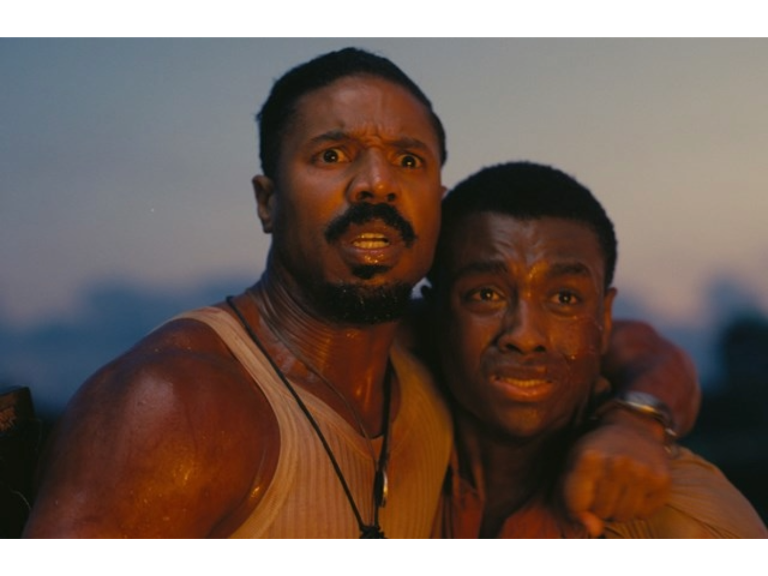 Two people standing close together outdoors during dusk, with one person’s arm around the other in a supportive gesture. Both are wearing sleeveless tops, and their skin appears glistening as if from sweat or water. The background shows a soft, blurred horizon with muted blue and orange tones, suggesting evening light. The image conveys a sense of physical exertion or intensity in a natural setting.