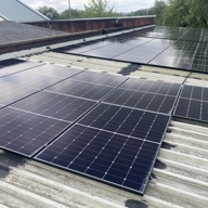 Rows of black solar panels installed on a corrugated metal rooftop. The panels are arranged in neat grids, reflecting light under a cloudy sky. In the background, there are additional rooftops, trees, and a glimpse of the horizon.