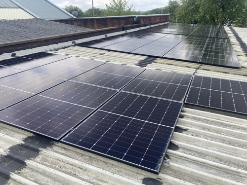 Rows of black solar panels installed on a corrugated metal rooftop, with additional panels visible further back. Surrounding elements include a brick wall, part of a sloped roof, and green trees in the background under a partly cloudy sky.