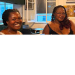 Two people sit indoors at a café or community venue, facing the camera. One wears a light-coloured sleeveless top and is smiling broadly, while the other wears glasses and a dark sleeveless top. Behind them are round tables, chairs, large windows with evening light outside, framed artwork on the walls, and a serving area or bar. The warmly lit space has a relaxed, welcoming atmosphere.