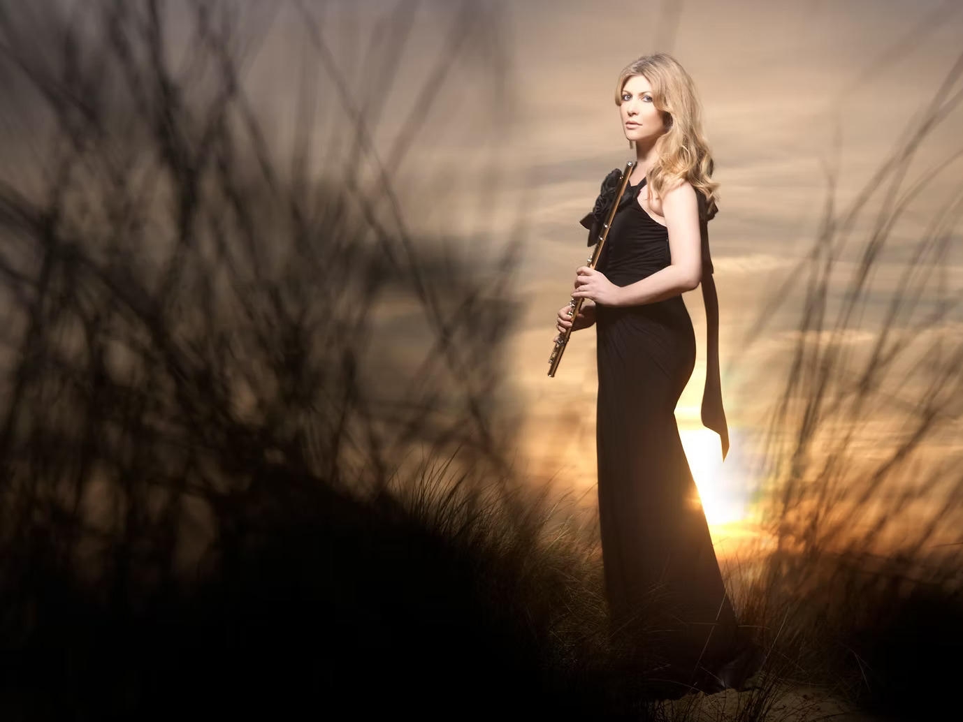 A woman in an evening dress stands in the countryside at sundown.