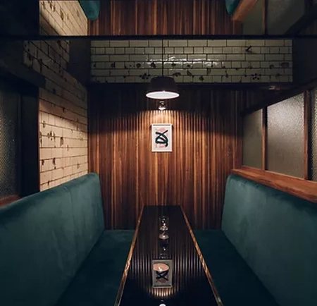 A cozy booth in a dimly lit bar or restaurant with teal upholstered seating on both sides of a long, glossy black table. The walls feature a mix of vertical wooden panels and white subway tiles with dark grout. A single pendant light hangs above the table, casting a warm glow. On the back wall, there is a framed minimalist artwork. The overall design combines modern and rustic elements, creating an intimate atmosphere.