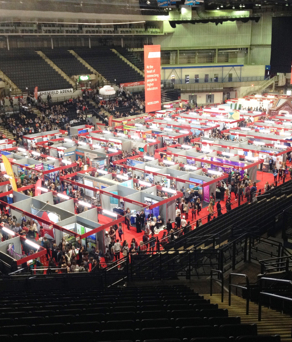 Large indoor exhibition at Sheffield Arena with rows of booths arranged on a red carpeted floor, surrounded by crowds of visitors. Elevated seating areas are visible in the background.