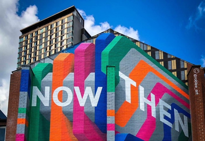 A vibrant mural on the side of a building displays the words 'NOW THEN' in large white letters against a colorful background of overlapping geometric shapes in blue, green, orange, pink, and purple. A tall building with many windows stands in the background under a partly cloudy sky. The artist's Instagram handle '@roblee.art' is visible on the right.