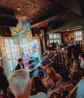  A crowded live music performance inside the Frog & Parrot pub, with warm lighting, patterned ceilings and a band playing near the front windows while people gather closely to watch.
