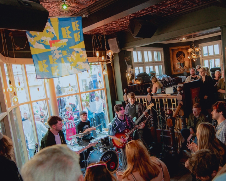  A crowded live music performance inside the Frog & Parrot pub, with warm lighting, patterned ceilings and a band playing near the front windows while people gather closely to watch.
