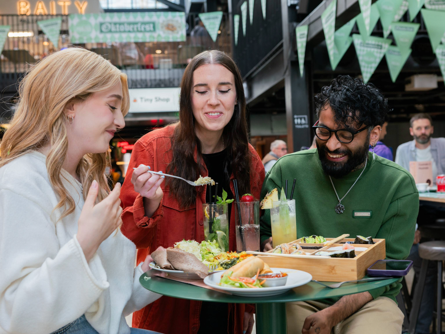Three people seated at a round green table in an indoor food hall, sharing a meal. The table holds plates of tacos, salad, bread, and a wooden tray with assorted dishes. There are also tall glasses of colourful drinks garnished with fruit and herbs. In the background, green triangular banners hang overhead, and signs for “Oktoberfest” and “Baity” are visible, creating a lively, casual dining atmosphere.