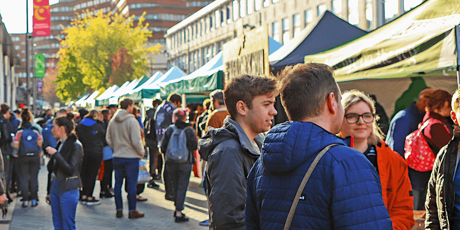 A busy outdoor vegan market with crowds browsing stalls lined along a city street, featuring colourful canopies and vendors serving food and handmade goods.