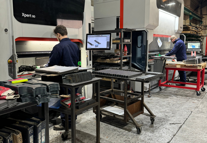 Industrial workshop with two large press brake machines labeled ‘Bystronic Xpert 80.’ Workstations with metal parts, tools, and a computer monitor displaying technical diagrams are positioned in front of the machines. Overhead beams and shelving with materials are visible in the background.