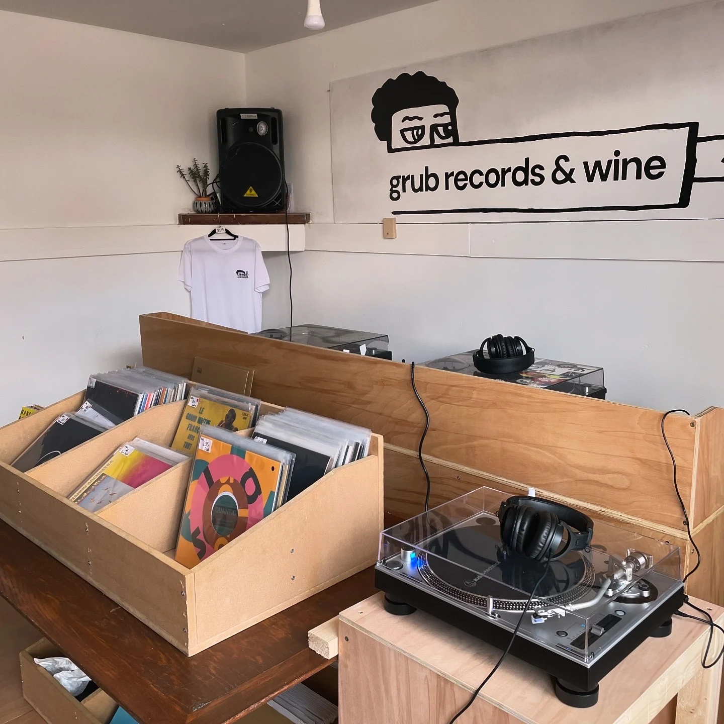 Interior of a small record shop with wooden bins filled with vinyl records, a turntable with headphones on the counter, and a wall sign reading ‘grub records & wine’ above a hanging white T-shirt.