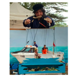 A puppeteer stands behind a small outdoor puppet theatre, manipulating strings attached to a model fishing boat and miniature figures. The scene features a white boat marked “YH3” floating on painted blue water, with puppet characters on board and alongside the vessel. A canopy tent and trees form the background, creating the setting for a live marionette performance.