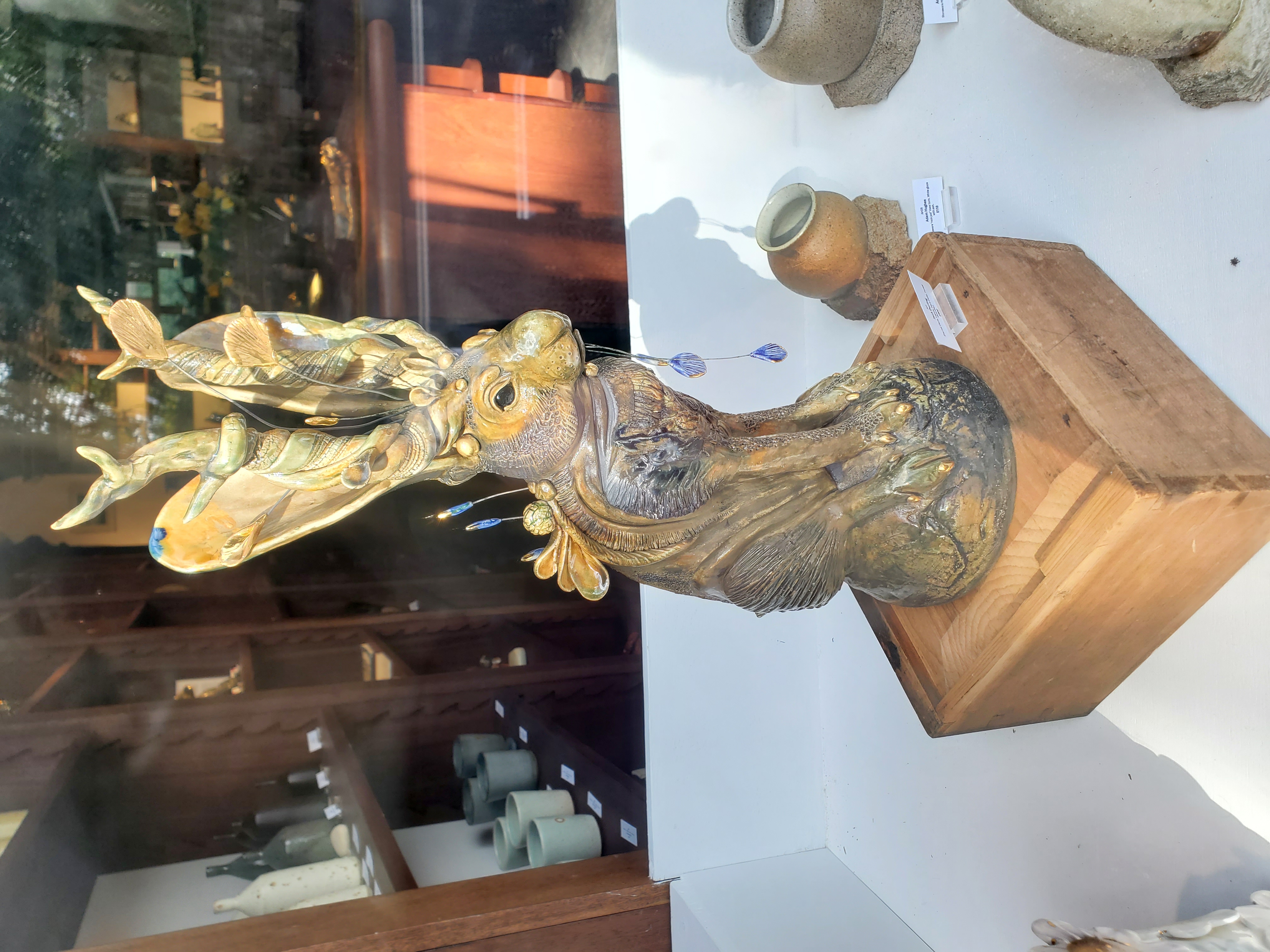 Decorative sculpture of a seated hare displayed in an art gallery window. The hare is finished in bronze and gold tones and features large, leaf-shaped ears adorned with miniature animal and plant forms. Blue decorative drops hang from the sculpture, while ceramic vessels and gallery shelving are visible in the background through the glass.