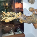 Decorative sculpture of a seated hare displayed in an art gallery window. The hare is finished in bronze and gold tones and features large, leaf-shaped ears adorned with miniature animal and plant forms. Blue decorative drops hang from the sculpture, while ceramic vessels and gallery shelving are visible in the background through the glass.