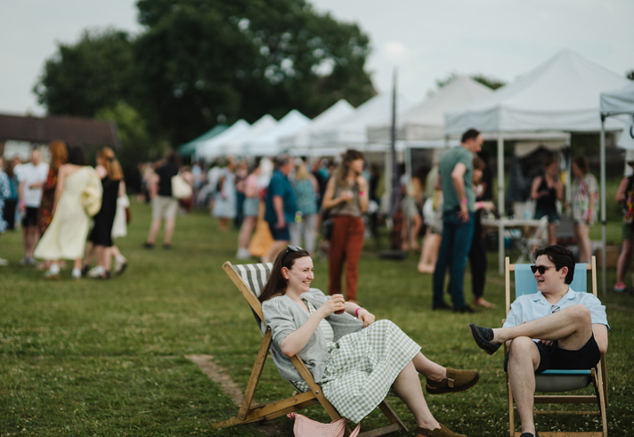 An outdoor event with lots of marquees, crowds of people walking about and some sitting in deck chairs.