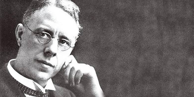 Black-and-white photograph of a person wearing a formal suit with a collared shirt and tie, seated and resting one arm on a surface while the other hand is positioned near the chin. The background is plain and dark, creating a classic portrait style.