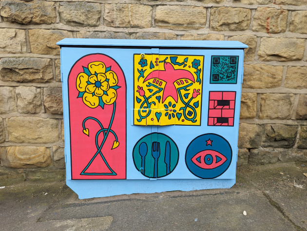 Utility box painted with colorful geometric artwork featuring a yellow flower on a pink background, stylized birds and hearts on a yellow panel, a QR code, brick pattern, and circular icons including cutlery and an eye symbol. The box is set against a stone wall.