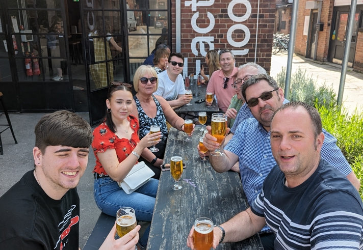 A group of people sat round a table, outside a restaurant, drinking beers.