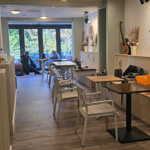 Interior of a cozy café with wooden flooring and light-colored walls decorated with dried floral arrangements and a mounted guitar. Several tables and chairs are arranged along the narrow space, with a bench running along the right wall. A small pumpkin decoration sits on a shelf to the left. Large glass doors at the back open to a view of trees outside, and a few people are seated near the windows.