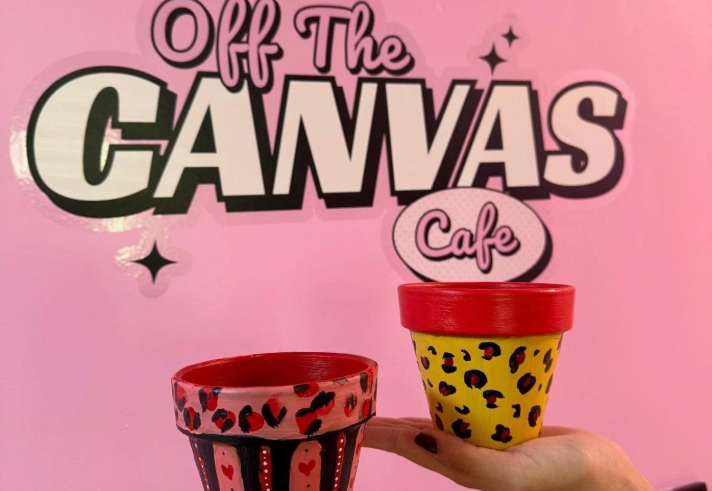 A hand is holding up a decorated pot in front of a  pink wall that has logo on it that reads 'OFF THE CANVAS'.
