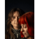 Portrait of two women against a dark background. One woman with bright red hair is in the foreground, shown in soft focus and looking downward, while a second woman with brown hair stands behind her, partially in focus and looking towards the camera. The image uses shallow depth of field and warm lighting to create an artistic, intimate composition.