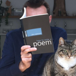 A person sits indoors at a table holding a book titled How to Write a Poem by John Redmond in front of their face. A tabby cat with green eyes sits in the foreground beside the book, looking directly at the camera. Shelves with plants, kitchenware and household items are visible in the background, creating a relaxed home setting. The image playfully contrasts the hidden reader with the attentive cat.