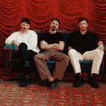 Three people sat on a sofa in front of a red pleated curtain.