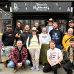 Group photo outside Abbeydale Beerworks Taproom. Thirteen people pose on the pavement and outdoor seating area in front of the venue’s black frontage and sign. Some are standing while others kneel at the front. Festoon lights hang across the shopfront, and neighbouring buildings are visible on either side. The image appears to have been taken during a gathering or opening event at the taproom.