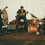 Three musicians perform live in an intimate indoor venue. A guitarist stands on the left, a bass player sits in the centre, and a drummer plays on the right. The stage area is decorated with warm white fairy lights hanging across the backdrop, creating a cosy atmosphere. Amplifiers, instruments and a patterned rug complete the setup, while soft lighting gives the performance a relaxed, live music event feel.