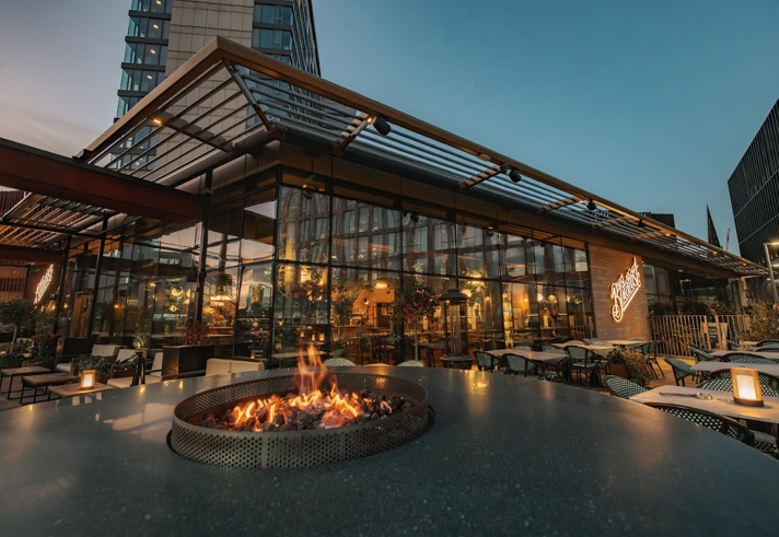 An evening view of a glass-walled restaurant surrounded by outdoor seating, with a circular fire pit lit in the foreground and soft lighting throughout the terrace.