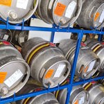 Rows of metal beer casks stored on blue metal shelving inside a warehouse. Each cask has an identification label attached.