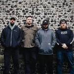 Four people stand side by side against a wall made of flint stones and brick. Dressed in casual jackets, hoodies and hats, they face the camera in an outdoor urban setting. The muted colours and textured stone backdrop create a simple, informal band-style group portrait.