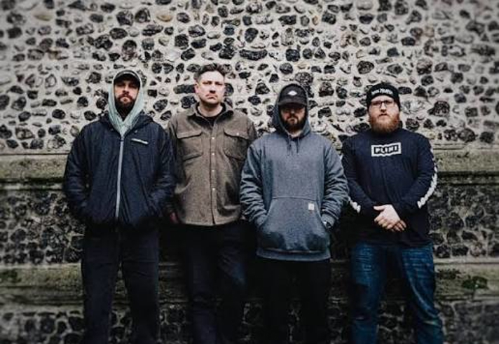 Four people stand side by side against a wall made of flint stones and brick. Dressed in casual jackets, hoodies and hats, they face the camera in an outdoor urban setting. The muted colours and textured stone backdrop create a simple, informal band-style group portrait.