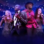 Four performers stand on stage holding microphones, dressed in sparkling and stylish outfits, including a sequined jacket and a bright red shirt. They are positioned in front of an ornate theatre interior with multiple balconies and glowing lights, creating a vibrant concert atmosphere with purple and blue stage lighting and mist in the foreground.