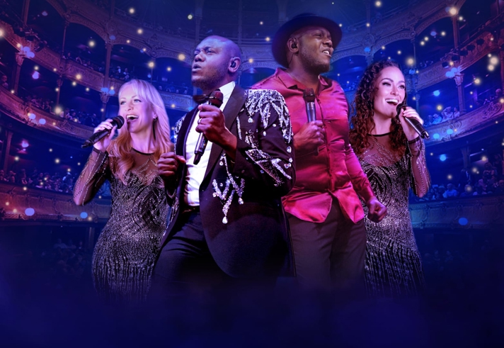 Four performers stand on stage holding microphones, dressed in sparkling and stylish outfits, including a sequined jacket and a bright red shirt. They are positioned in front of an ornate theatre interior with multiple balconies and glowing lights, creating a vibrant concert atmosphere with purple and blue stage lighting and mist in the foreground.