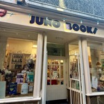 The exterior of an independent bookshop called “Juno Books,” featuring large front windows filled with posters and displays. The shopfront is cream-coloured with a central doorway leading inside, and a string of turquoise fairy lights hangs above the entrance.